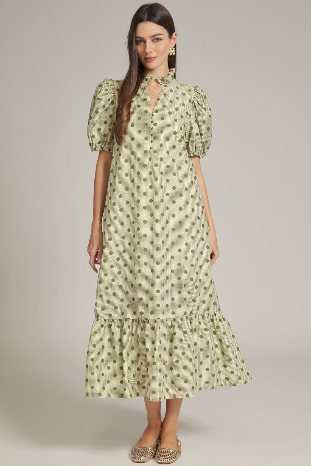 A woman modeling a light sage green midi dress with a dark green polka dot pattern, puff sleeves, and a tiered ruffle hem, set against a plain light gray studio background.