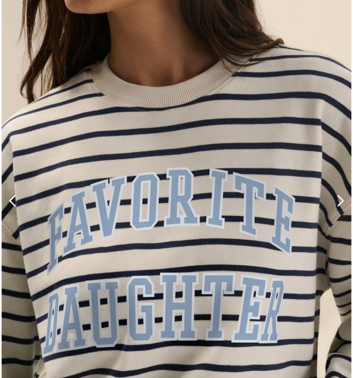 A close-up of a person wearing a cream-colored crewneck sweatshirt with navy blue horizontal stripes and light blue collegiate-style text reading 'FAVORITE DAUGHTER' across the front.