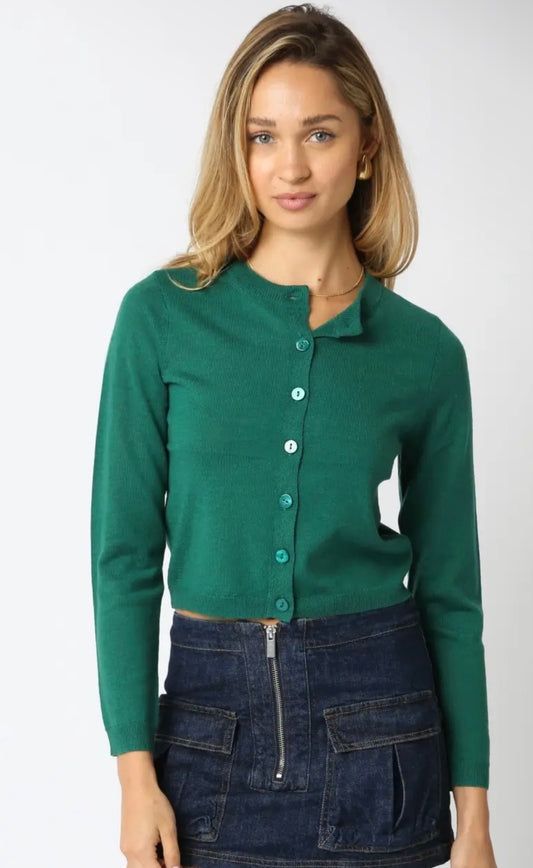 A woman wearing a green long-sleeve button-front cardigan top and a dark denim zip-front skirt, photographed against a plain light gray studio background.