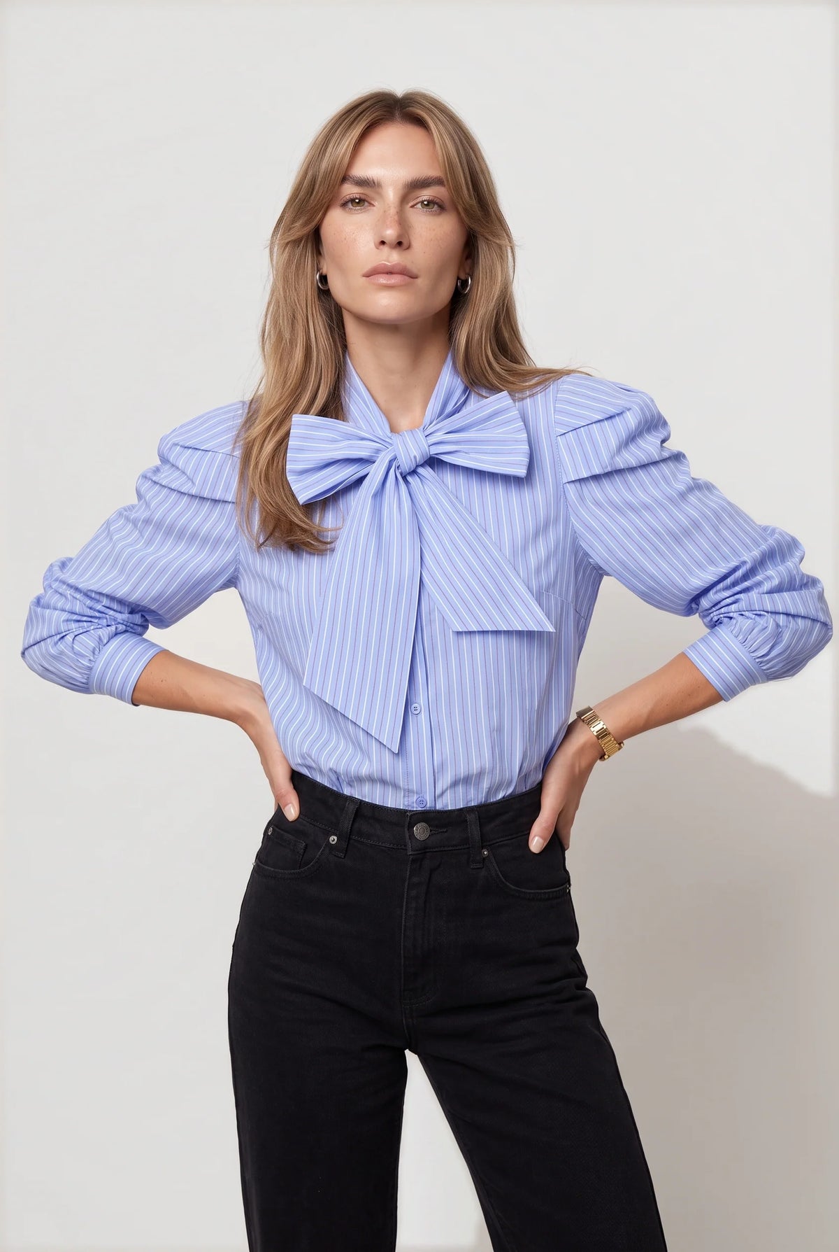 A woman modeling a light blue and white vertically striped blouse with a large bow at the collar, paired with black jeans, against a plain light gray studio background.