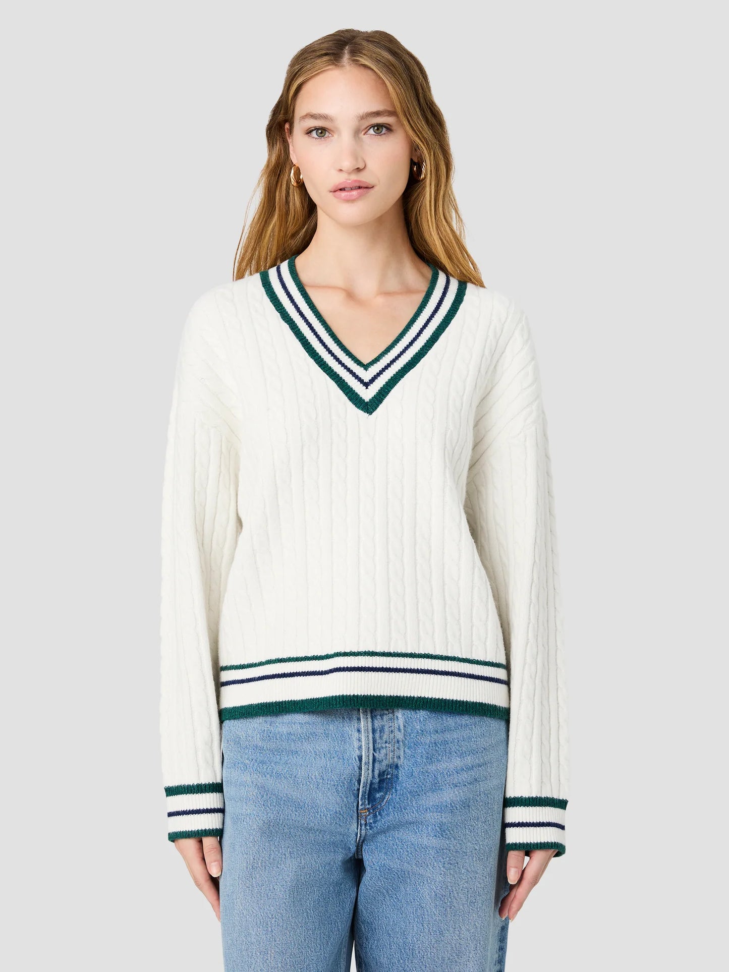 A woman modeling a cream ribbed knit sweater with a green and navy striped V-neck and hem, paired with light blue jeans, against a solid light gray background.