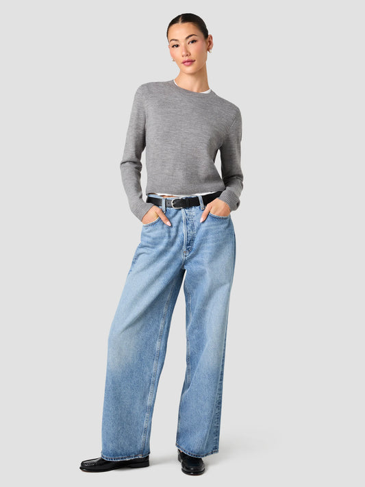 A model wearing a gray long-sleeve crewneck sweater, light-wash wide-leg blue jeans, a black belt, and black leather shoes, standing against a solid light gray background.