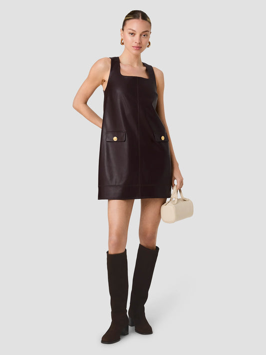 A model wearing a dark brown faux-leather mini dress with square neckline and gold buttons, paired with black knee-high boots and holding a cream-colored handbag, set against a solid light gray background.