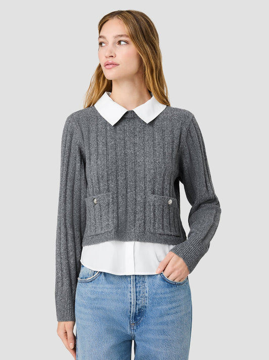A woman wearing a gray ribbed knit cropped cardigan with a white pointed collar, layered over a white blouse and blue jeans, against a solid light gray background.