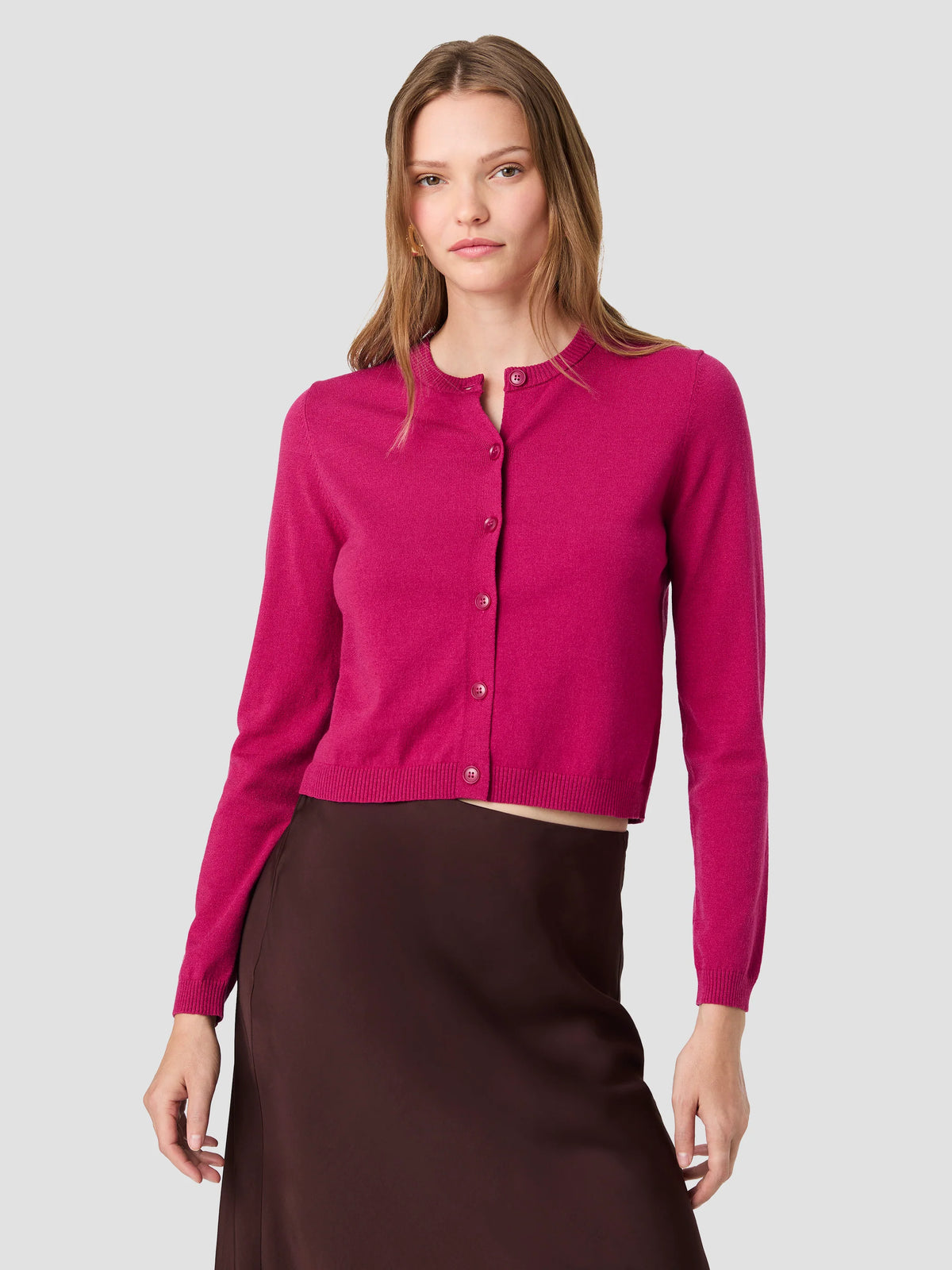 A woman modeling a bright magenta pink cropped button-up cardigan sweater paired with a dark brown skirt, set against a solid light gray background.