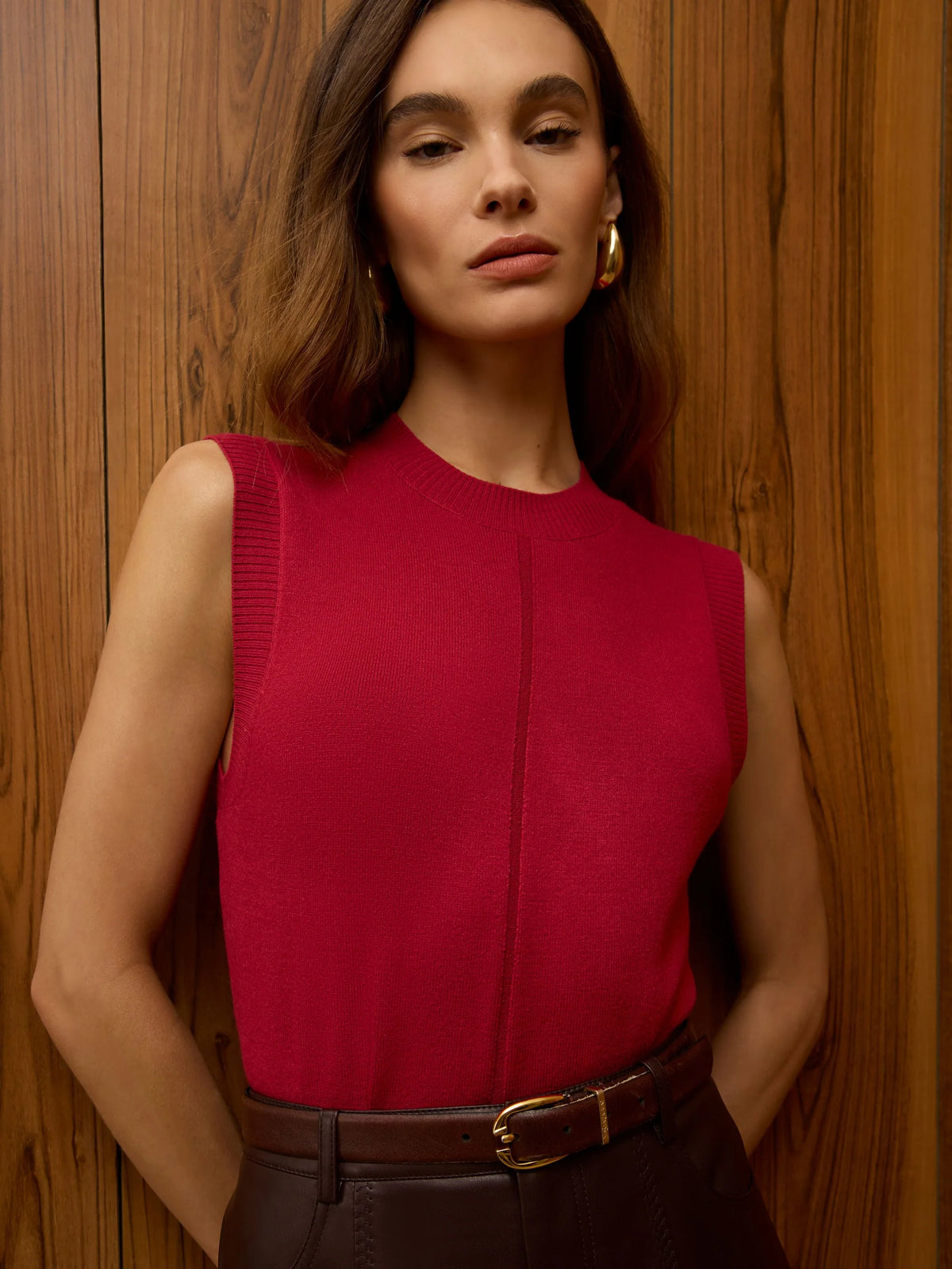 A woman wearing a sleeveless, crew-neck knit top in a rich magenta-red color, paired with a brown belt and dark brown trousers, standing against a warm wood-paneled wall.