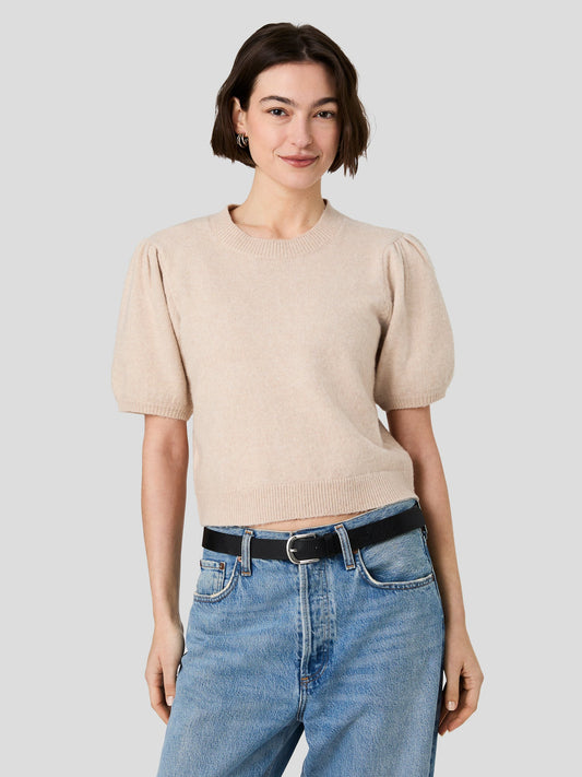 A woman modeling a light beige short-sleeve knit top with puffed sleeves, paired with blue jeans and a black belt, against a solid light gray background.