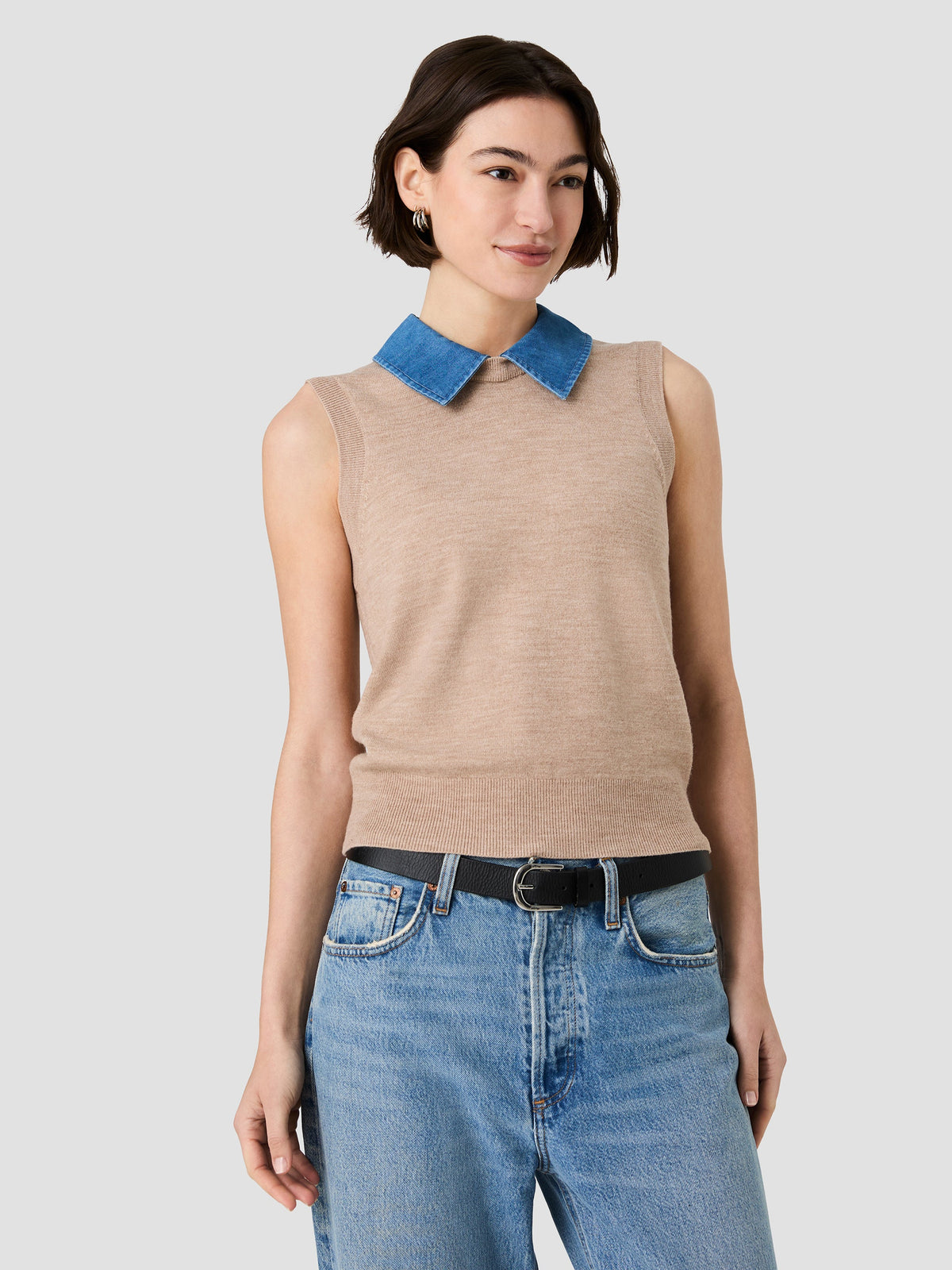 A woman modeling a sleeveless beige knit top with a contrasting blue collar, paired with blue jeans and a black belt, against a light gray studio background.