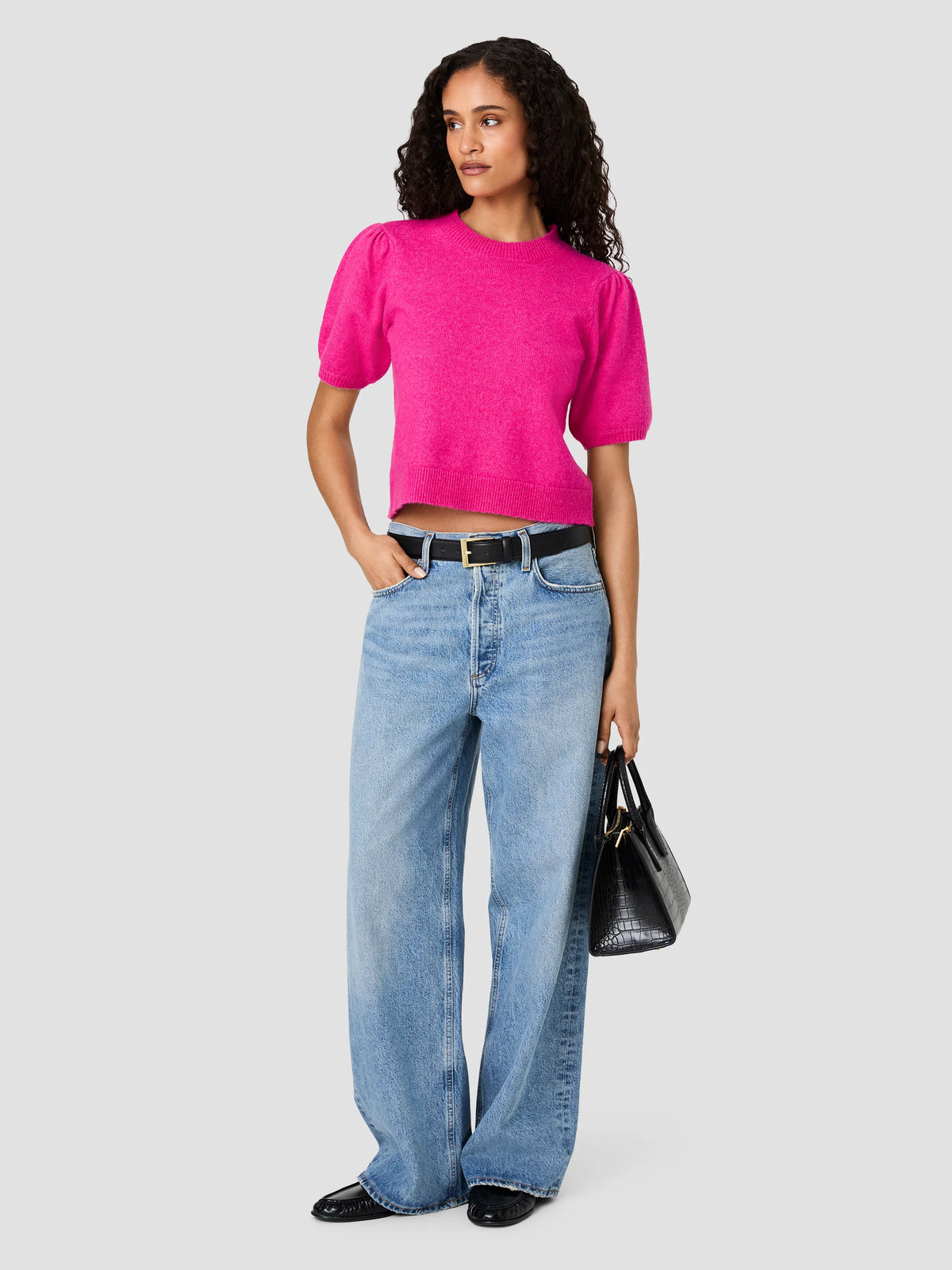 A model wearing a bright pink short-sleeve cropped knit top, light-wash wide-leg blue jeans, a black belt, and black shoes, holding a black handbag against a solid light gray background.
