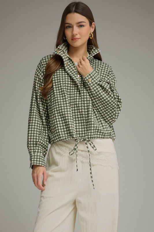 A woman modeling a green and white gingham long-sleeve shirt with a high collar and drawstring, paired with cream-colored pants, against a solid gray studio background.