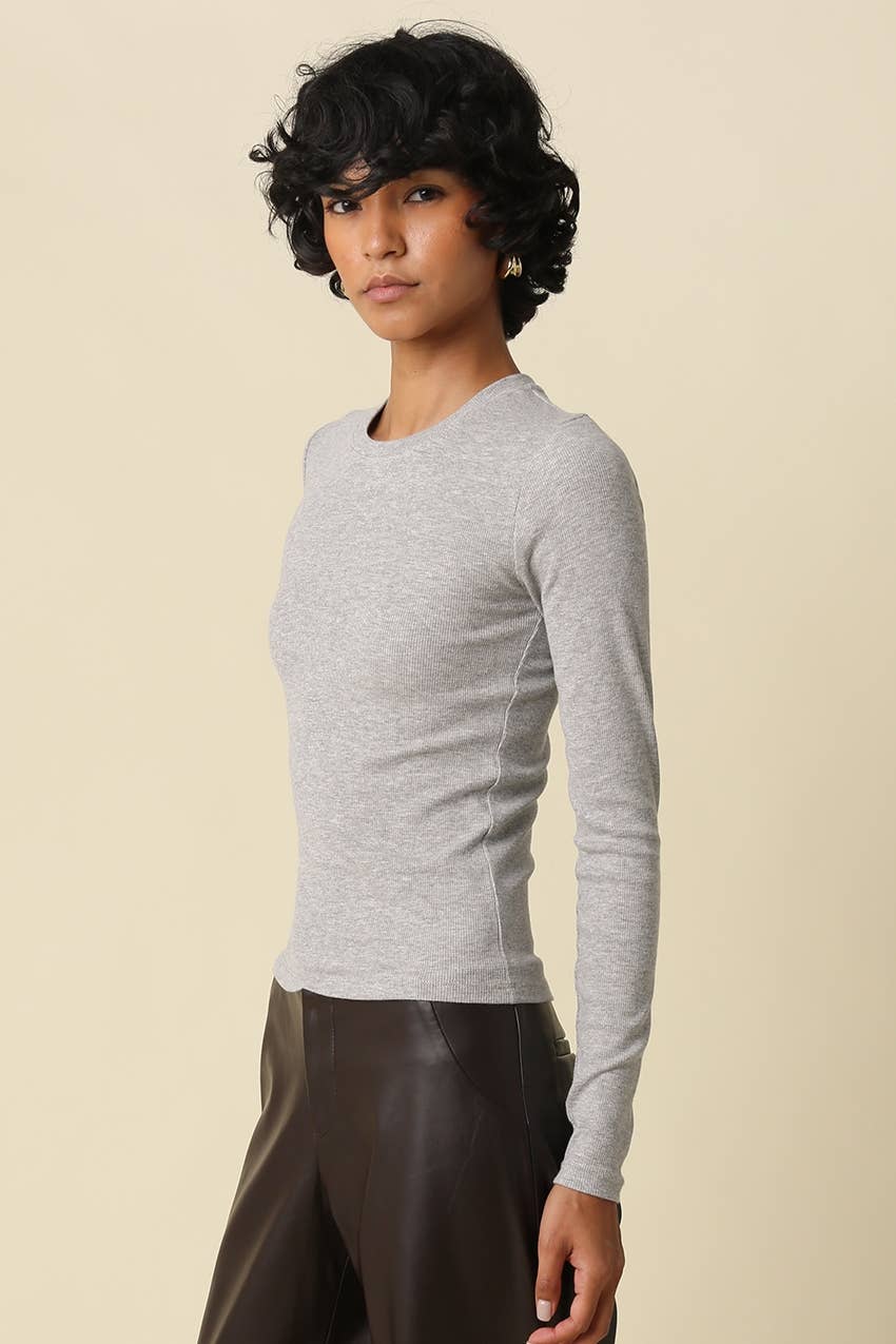 A model with short curly black hair wearing a light gray long-sleeve crewneck top and dark brown pants, standing against a solid beige background.