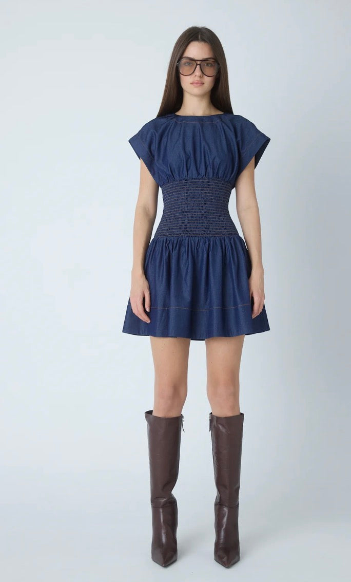 A woman modeling a navy blue short dress with cap sleeves and a smocked waist, paired with dark brown knee-high boots, against a light gray studio background.