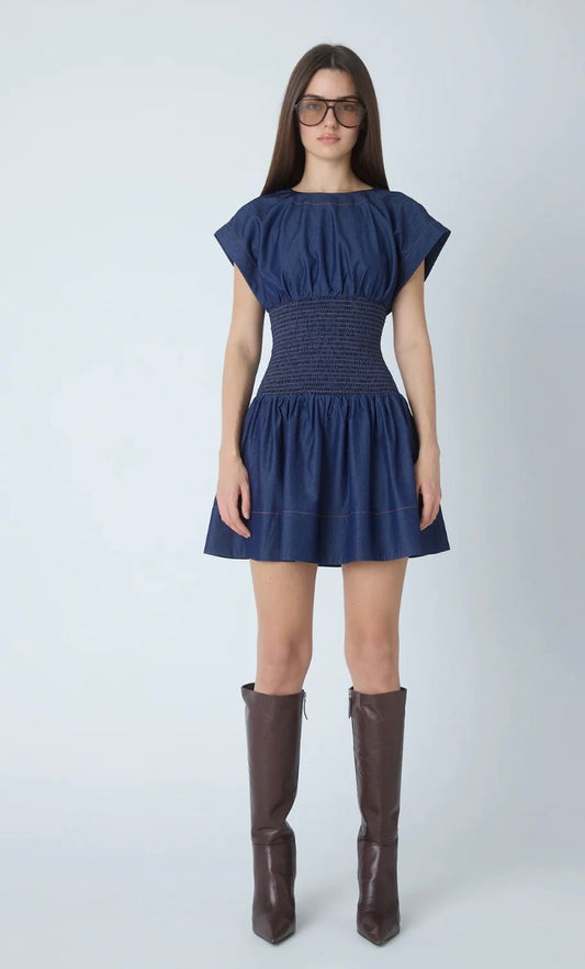 A woman modeling a navy blue short dress with cap sleeves and a smocked waist, paired with dark brown knee-high boots, against a light gray studio background.
