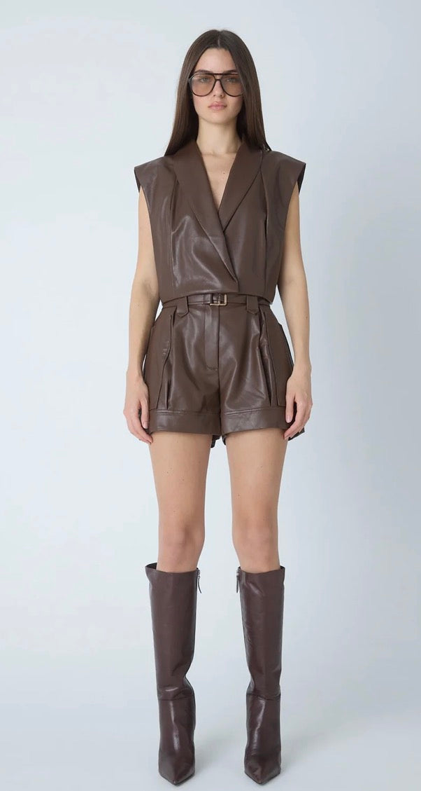 A model wearing a brown faux-leather sleeveless wrap vest and matching shorts with a belt, paired with brown knee-high boots, against a light gray studio background.