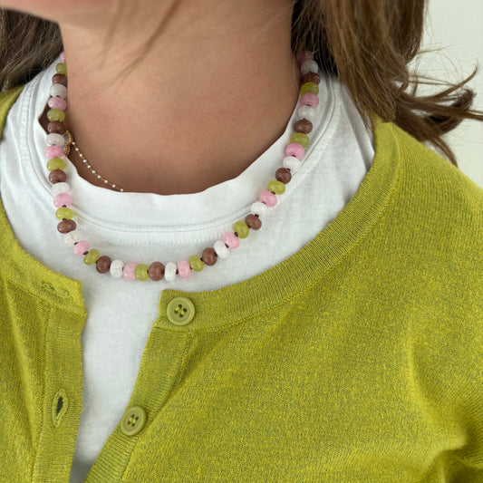 A close-up of a person wearing a beaded necklace with pastel pink, olive green, and brown beads over a white shirt and yellow-green cardigan.