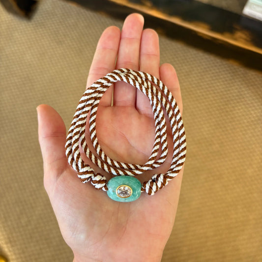 A close-up of a hand holding a braided rope bracelet with a turquoise bead and a small gold-toned circular charm in the center, set against a beige textured background.