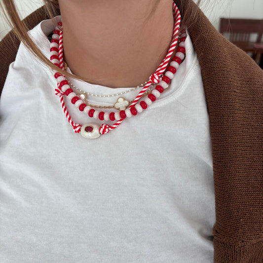 A close-up of a person wearing a layered necklace set. The necklace features red and white striped braided cords, red and white beads, a small white flower charm, a delicate white beaded strand, and a white circular charm with a red stone.