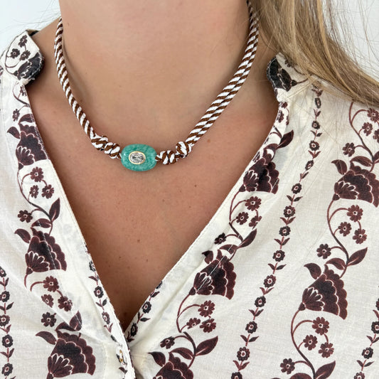 A close-up of a person wearing a braided rope necklace with a turquoise-colored oval pendant featuring a silver-toned circular center, paired with a cream blouse with a dark floral pattern.