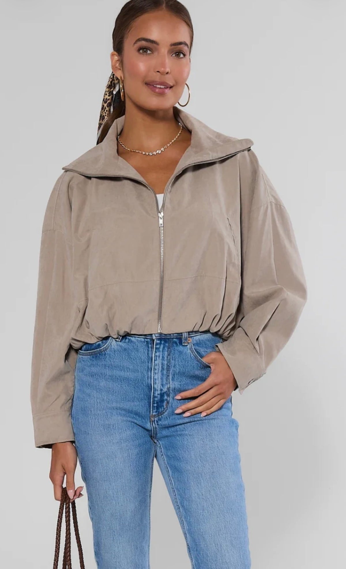A woman modeling a cropped beige zip-up jacket with a high collar, paired with blue jeans, against a light gray studio background.