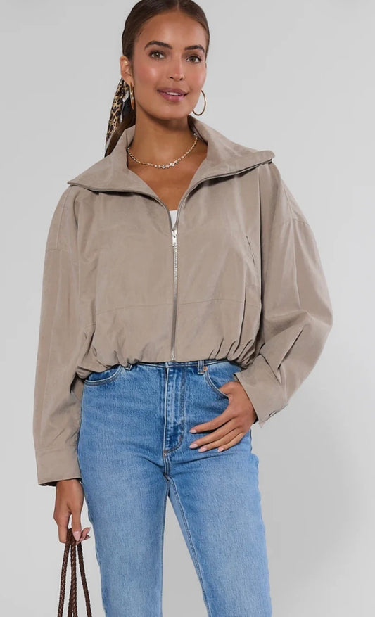 A woman modeling a cropped beige zip-up jacket with a high collar, paired with blue jeans, against a light gray studio background.
