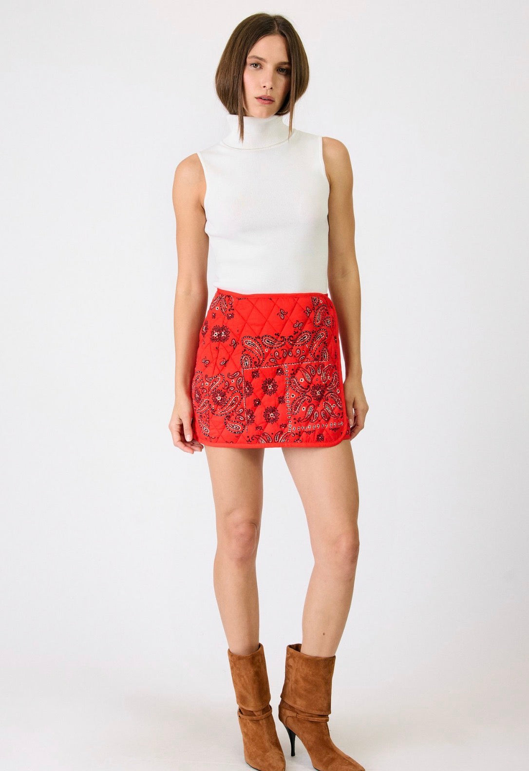 A model wearing a white sleeveless mock-neck top and a red mini skirt with a black paisley pattern, paired with brown suede ankle boots, against a plain white studio background.