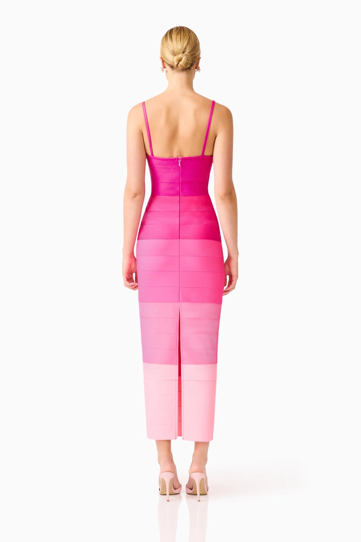 A woman viewed from the back wearing a sleeveless midi dress with a color-blocked design in shades of magenta, hot pink, and light pink, paired with nude high heels against a white studio background.