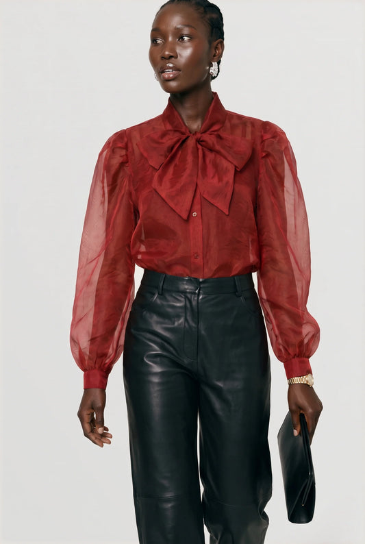 A model wearing a red sheer long-sleeve blouse with a large bow at the collar, paired with black leather pants, holding a black clutch against a light gray studio background.