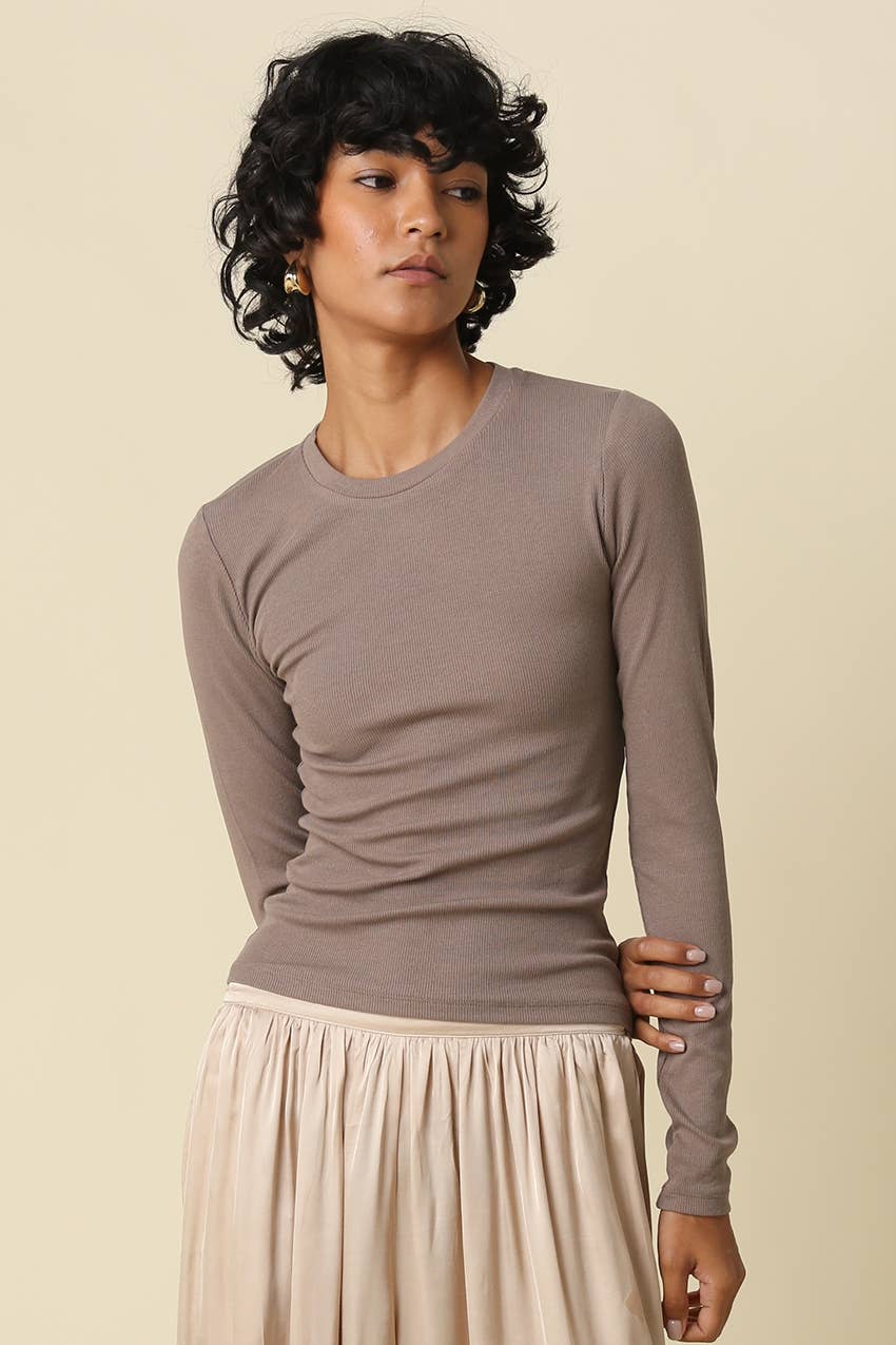 A woman modeling a fitted, long-sleeve crewneck top in taupe, paired with a pleated cream skirt, against a solid beige background.