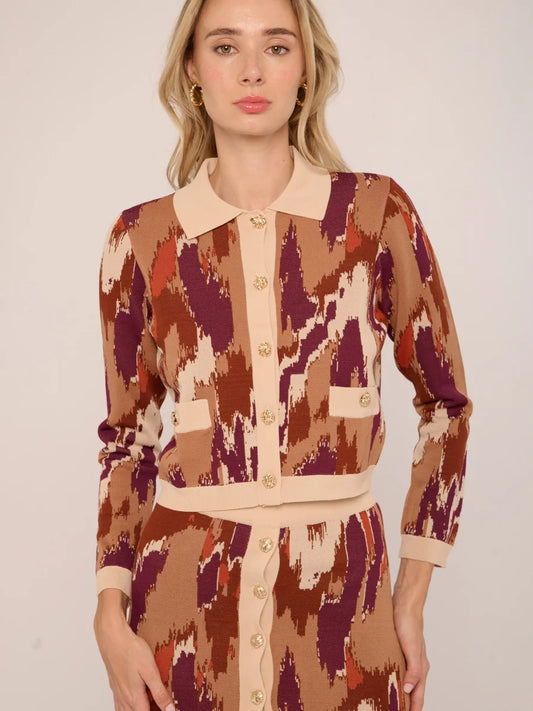 A woman modeling a matching long-sleeve button-front cardigan and skirt set with an abstract ikat-style pattern in tan, burgundy, rust orange, and cream, against a plain light beige studio background.