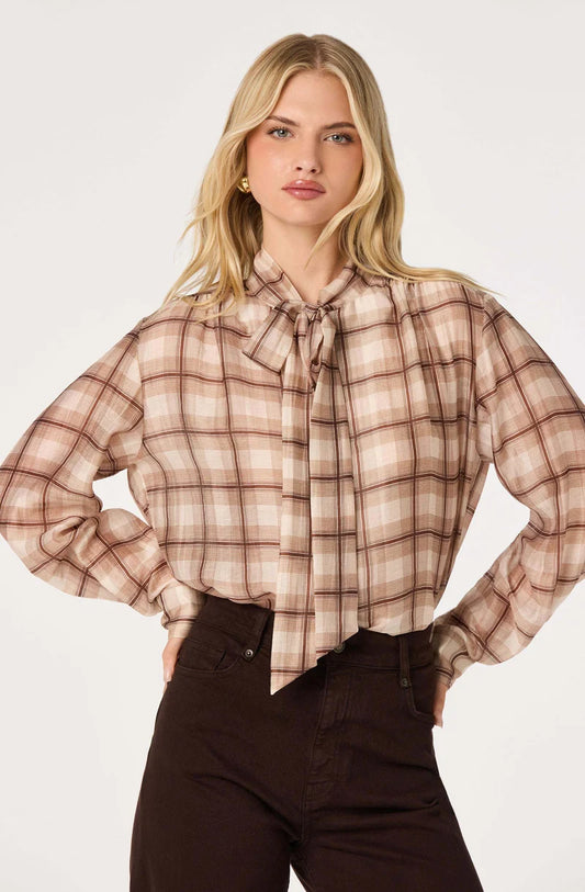 A woman modeling a long-sleeve plaid blouse with a bow tie neckline, paired with dark brown pants, against a plain light gray studio background.