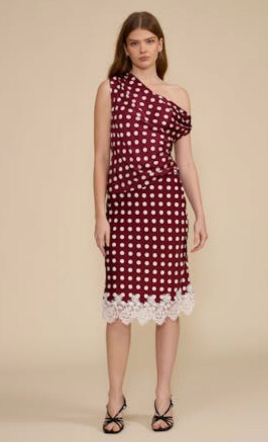 A model wearing a one-shoulder midi dress in burgundy with white polka dots and a white lace hem, paired with black strappy heels, against a solid beige background.