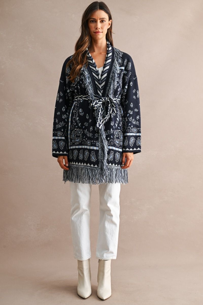 A woman stands against a plain beige studio background wearing a dark navy patterned wrap-style cardigan with fringe, white pants, and white ankle boots.