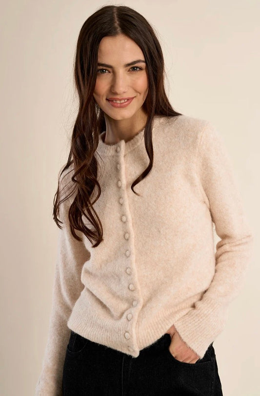 A woman with long dark brown hair wearing a fuzzy, light beige button-up cardigan sweater and black pants, posing against a solid light beige background.