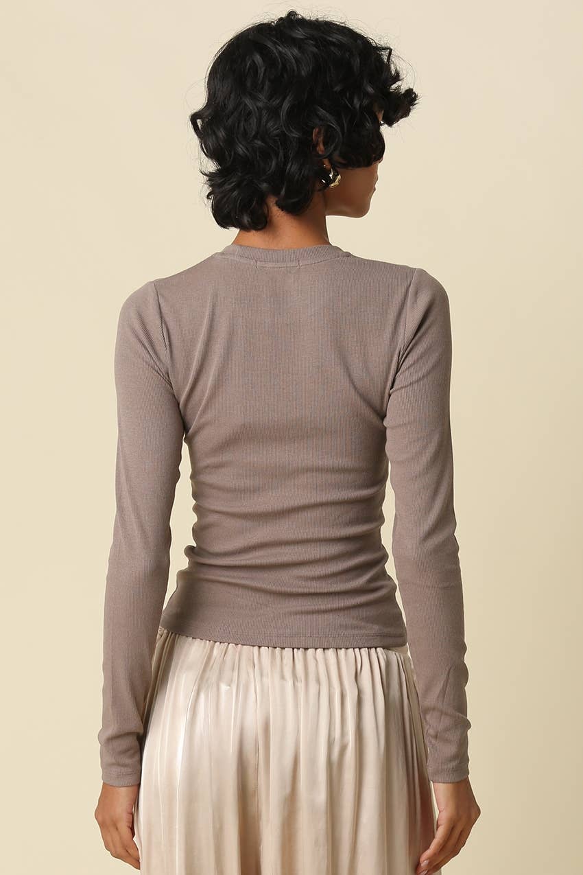 A woman shown from the back wearing a fitted, long-sleeve crewneck top in a muted taupe-brown color, paired with a pleated cream skirt, against a solid light beige background.