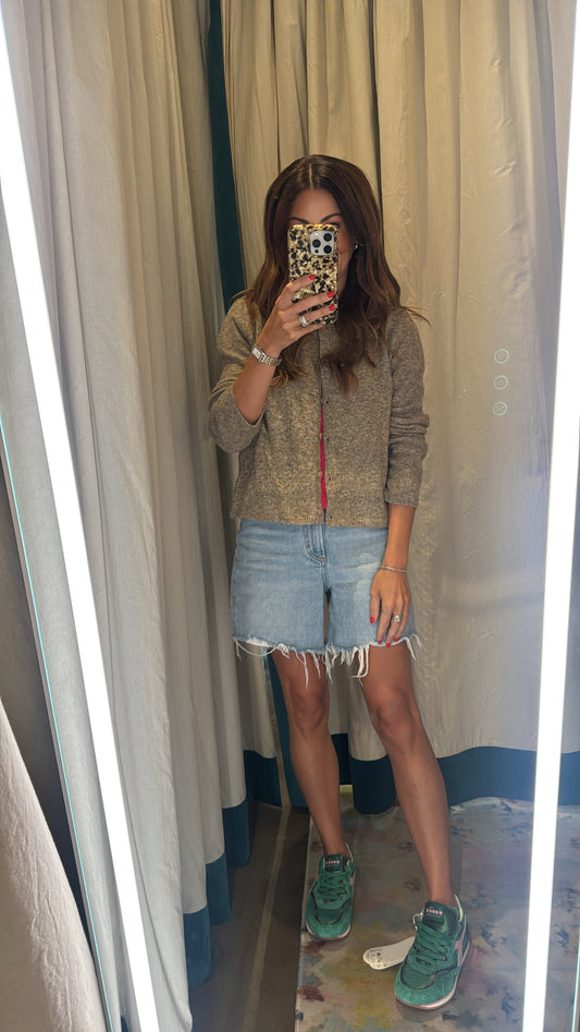 A woman takes a mirror selfie in a fitting room wearing a brown knit zip-up hoodie, a light-wash denim mini skirt with raw hems, and green sneakers.