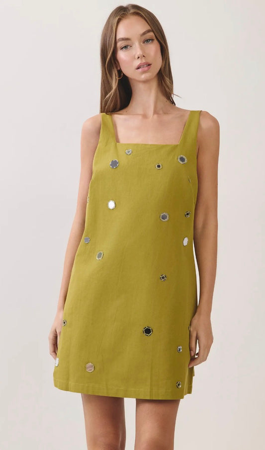 A woman modeling a sleeveless olive yellow dress with square neckline and scattered silver and black circular embellishments, set against a plain light gray studio background.