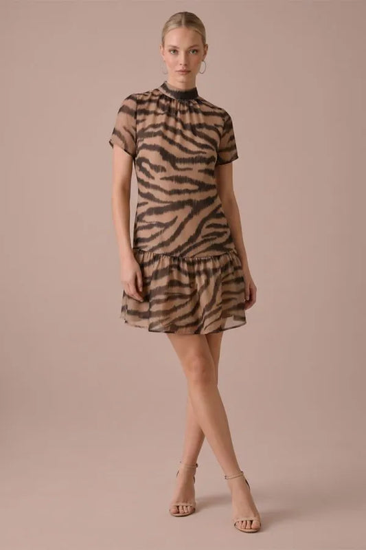 A woman modeling a short-sleeve tiered mini dress with a brown and beige zebra print, paired with beige high-heeled sandals, against a solid dusty pink studio background.