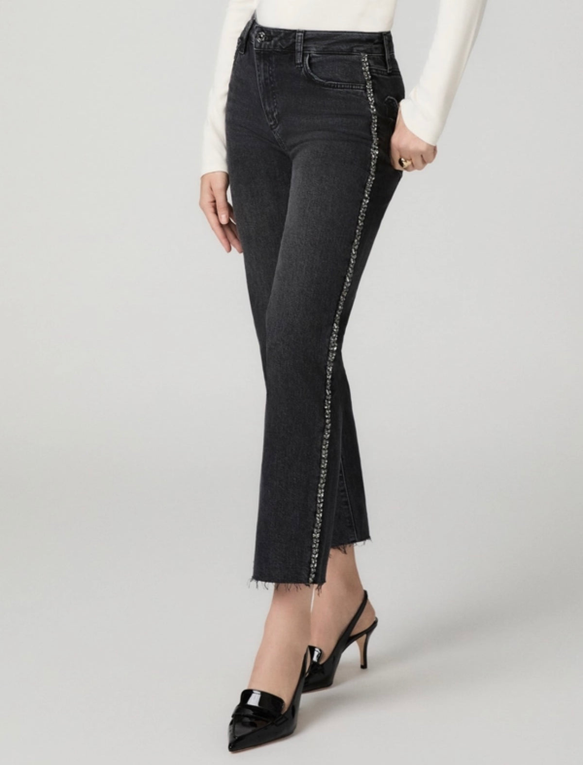 A model wearing dark gray flare jeans with a silver chain detail running down the side, paired with a white long-sleeve top and black slingback heels, against a light gray studio background.