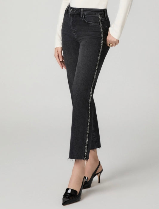 A model wearing dark gray flare jeans with a silver chain detail running down the side, paired with a white long-sleeve top and black slingback heels, against a light gray studio background.
