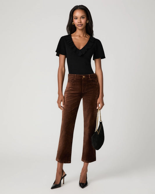 A model stands against a light gray studio background wearing a black short-sleeve ruffled top and brown cropped corduroy pants, holding a black handbag with a gold chain strap.