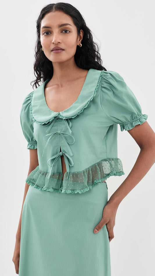 A woman modeling a mint green ruffled blouse with puff sleeves and a matching skirt against a plain light gray studio background.