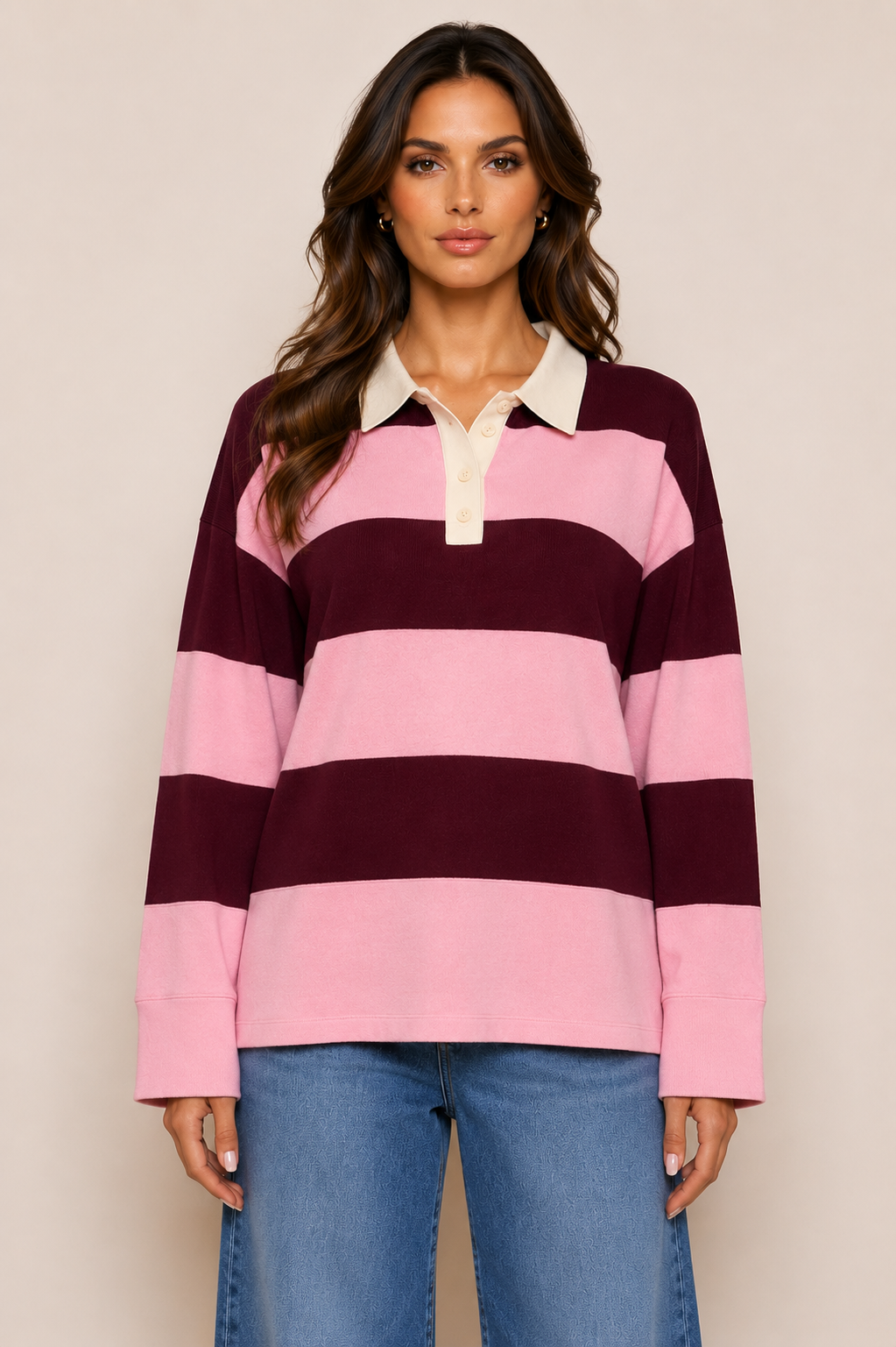 A woman modeling a long-sleeve rugby-style polo shirt with horizontal stripes in light pink and dark burgundy, featuring a cream-colored collar and placket, paired with blue jeans against a plain beige studio background.