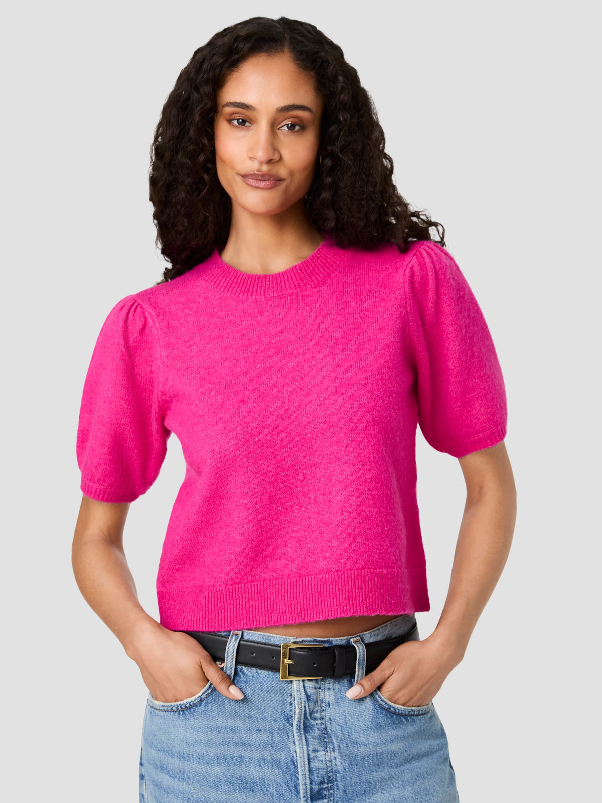 A woman modeling a bright pink short-sleeve knit sweater with puffed shoulders, paired with blue jeans and a black belt, against a solid light gray background.