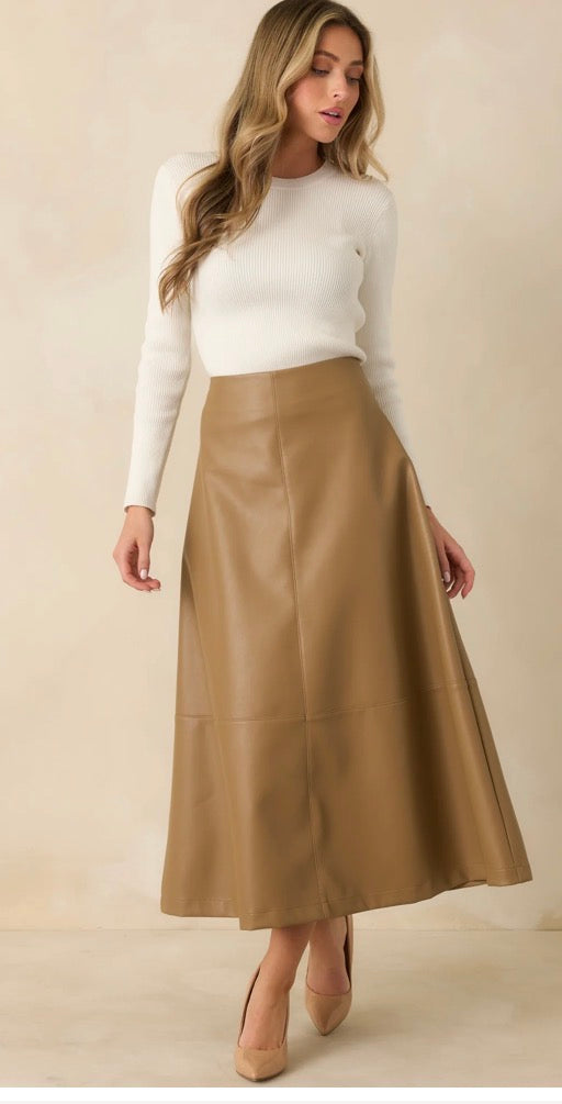 A woman modeling a high-waisted, A-line midi skirt in tan faux leather, paired with a white long-sleeve knit top and beige pointed-toe heels, against a warm beige studio background.