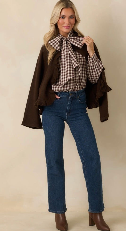 A woman modeling a brown ruffled cape-style jacket over a brown and beige gingham blouse with a large bow, paired with dark blue jeans and brown heeled boots, against a plain beige studio background.