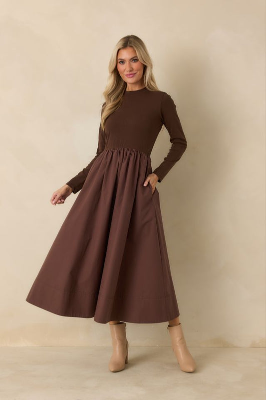 A woman modeling a long-sleeve brown midi dress with a gathered waist and side pockets, paired with beige ankle boots, against a neutral beige studio backdrop.