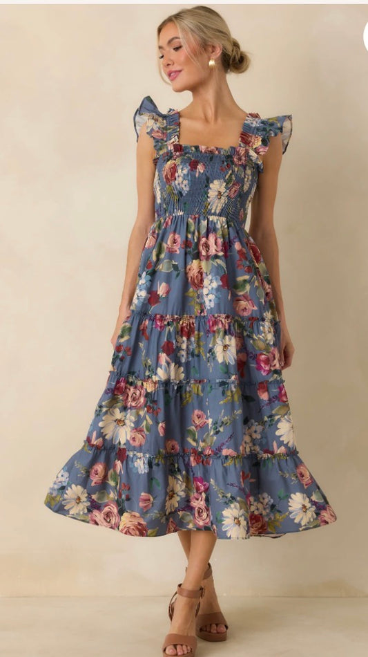 A woman modeling a tiered midi dress with a blue floral print, ruffled cap sleeves, and a square neckline, set against a plain beige studio background.