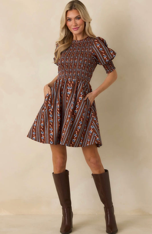 A woman modeling a short puff-sleeve fit-and-flare dress with an all-over brown, beige, and blue geometric pattern, paired with dark brown knee-high boots, against a solid beige studio background.