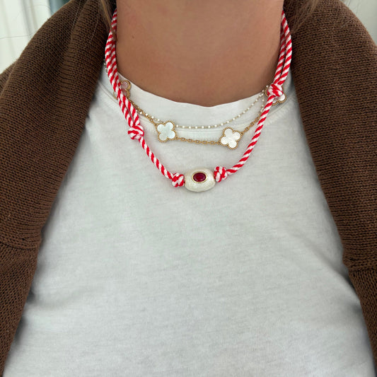 A close-up of a person wearing a layered necklace set. The set includes a red and white braided cord necklace with a silver-tone oval medallion featuring a red center, a gold-tone chain with three white flower charms, a pearl-like chain with a white flower charm, and a silver-tone chain with a white flower charm.