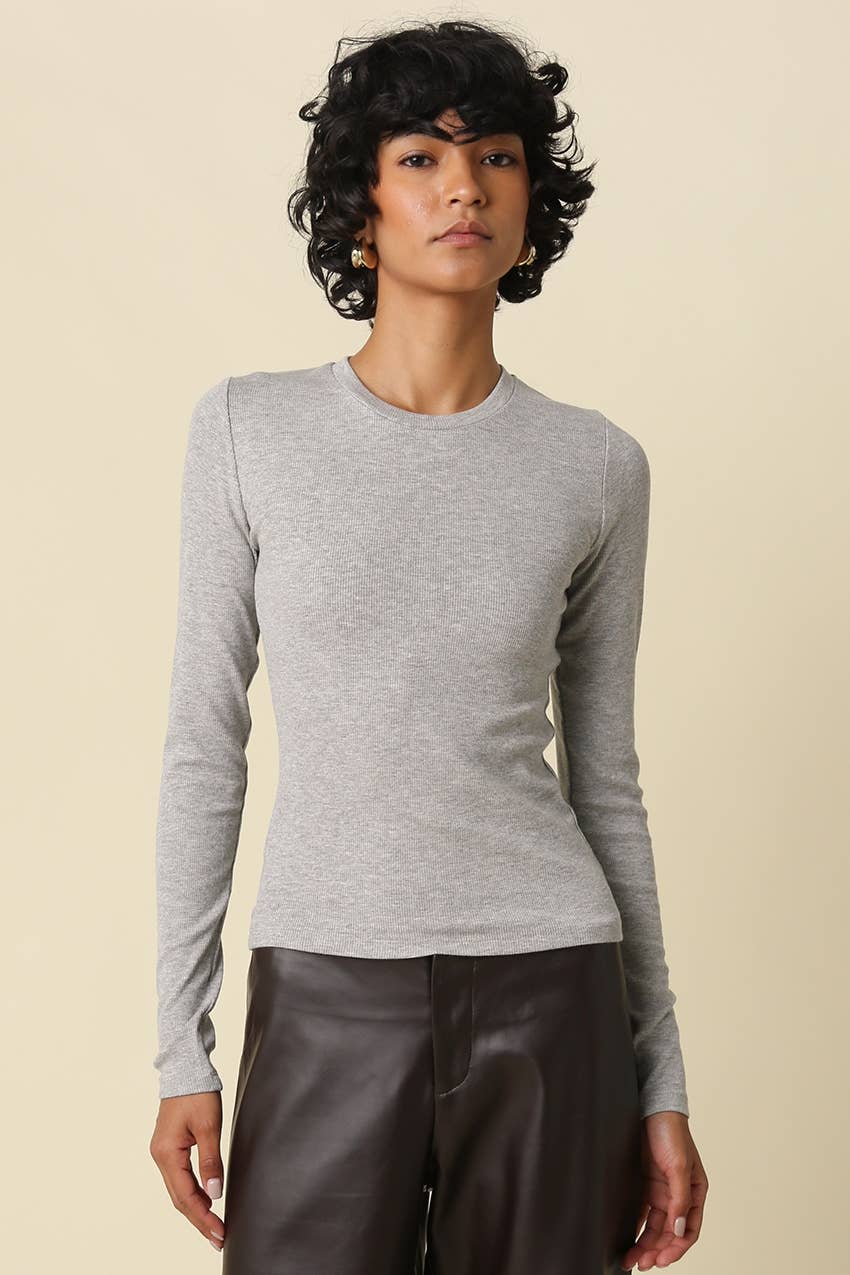 A woman modeling a light gray long-sleeve crewneck knit top with structured shoulders, paired with dark brown leather pants, against a solid beige background.
